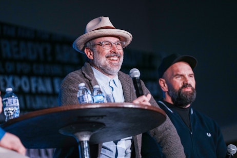 The image features Jason Lee and Ethan Suplee sitting together, likely on a panel at a fan or cultural event. Jason is on the left, wearing a hat and glasses, smiling, and holding a microphone, suggesting he is amid a discussion or presentation. His attire, including a tweed jacket and a tie, gives him a scholarly or artistic demeanor. Ethan is on the right, with a beard and a cap, has a more serious expression, and is also dressed casually. The background is dimly lit, focusing the viewer's attention on the speakers. The presence of water bottles on the table and microphones suggests a formal yet relaxed speaking event, such as a Q&A session, seminar, or live interview at a convention.