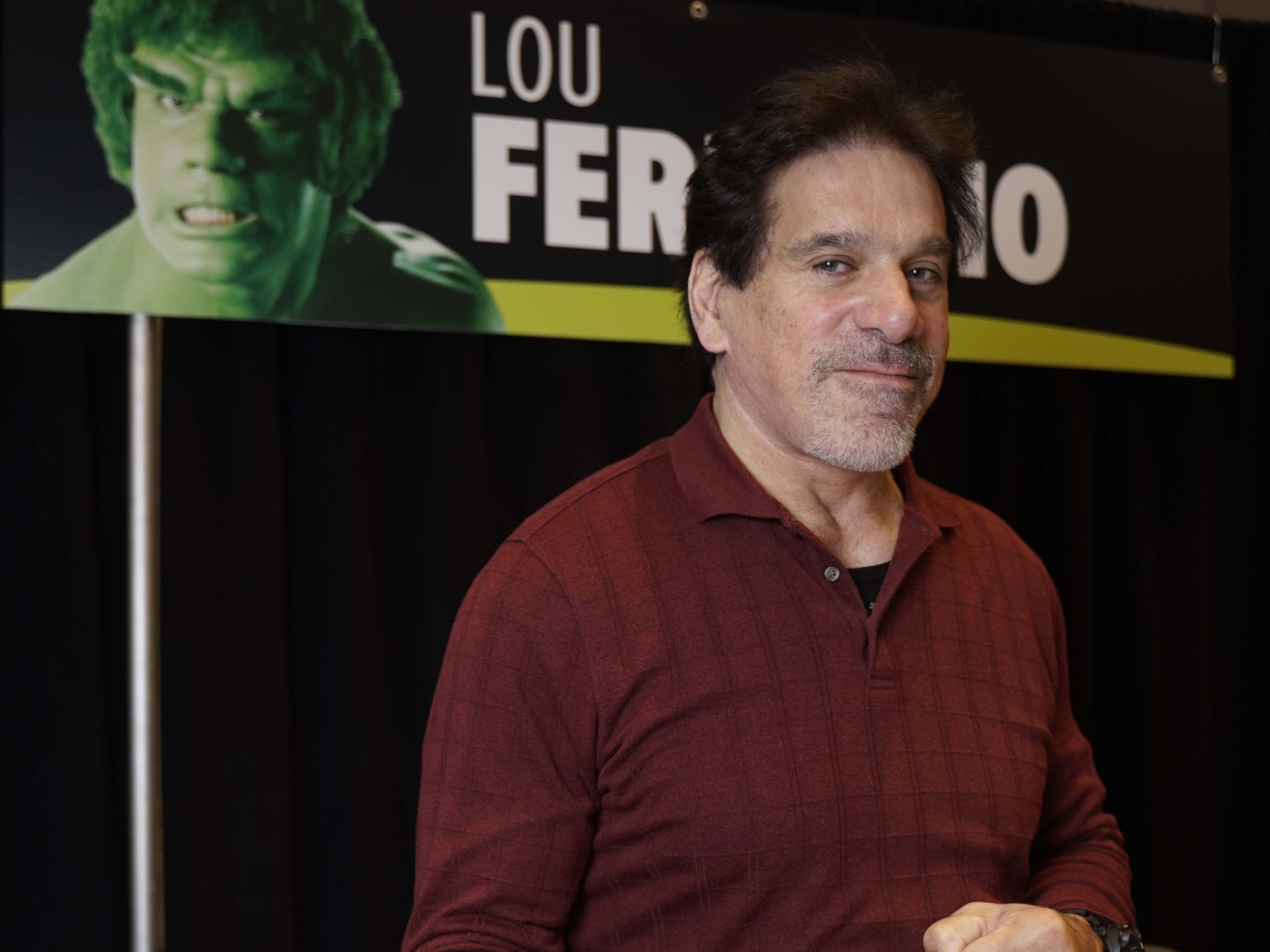 Lou Ferrigno doing an Incredible Hulk pose infront of a banner with the Hulk on it