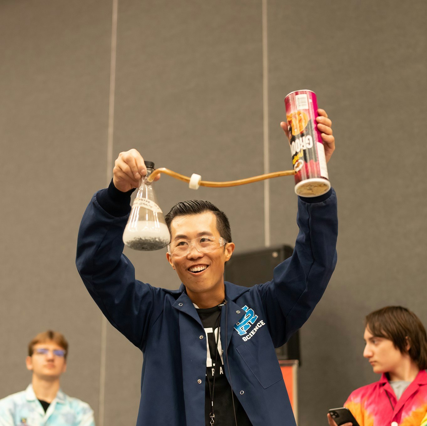 Man in lab coat with protective glasses. He is holing a scientific flask with a tube attached to a pringles can.