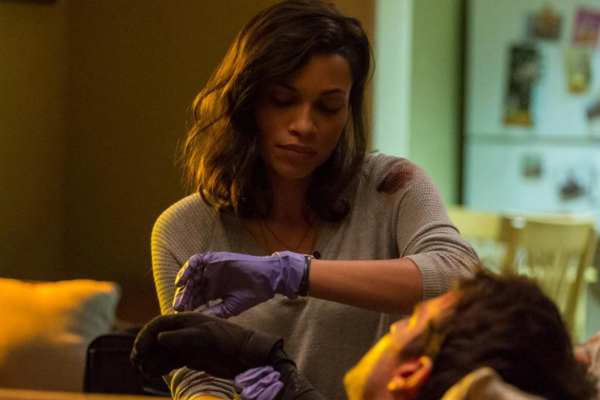 Rosario Dawson in a gray shirt with a blood stain on her shoulder, wearing surgical gloves. She is looking at her watch and taking Matt Murdock's pulse.