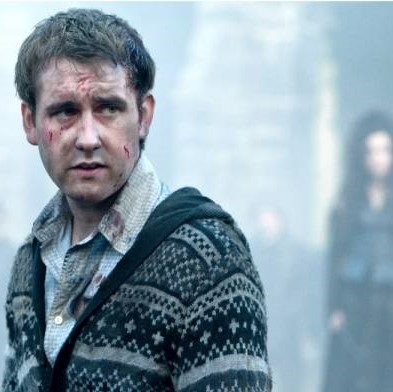 Nellille is wearing a sweater with a button up shirt underneath. He has dirt and cuts all over his face. In the background you can make out Bellatrix