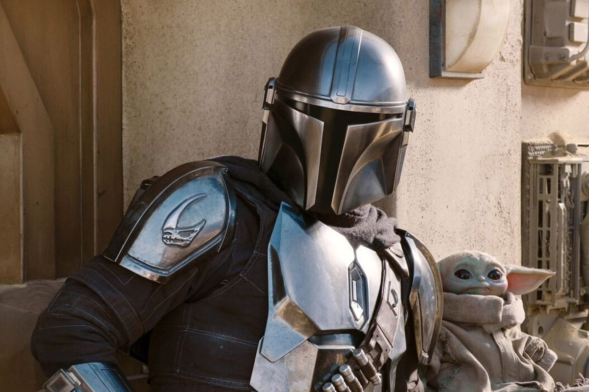 The Mandalorian stands against a beige wall holding Grogu in his arms