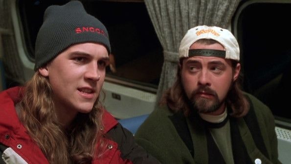 Jay and Silent Bob on a bus. Jay is wearing a red jacket and a black hat that said Snoochie. Silent Bob is looking at him, wearing his iconic backwards hat, dark green coat on top of the layer of shirts.