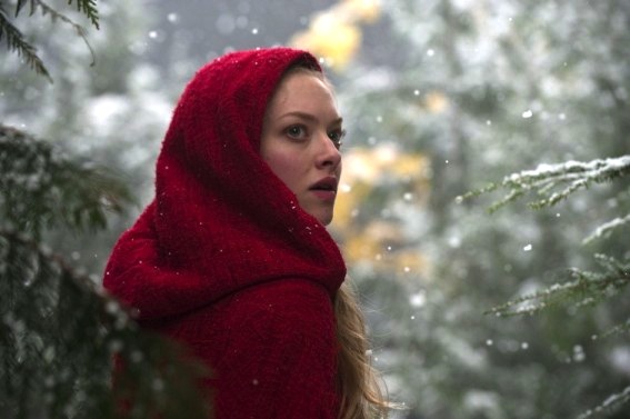A woman with blonde hair is wearing a red hood and is looking out into the distance in a snowy forest.