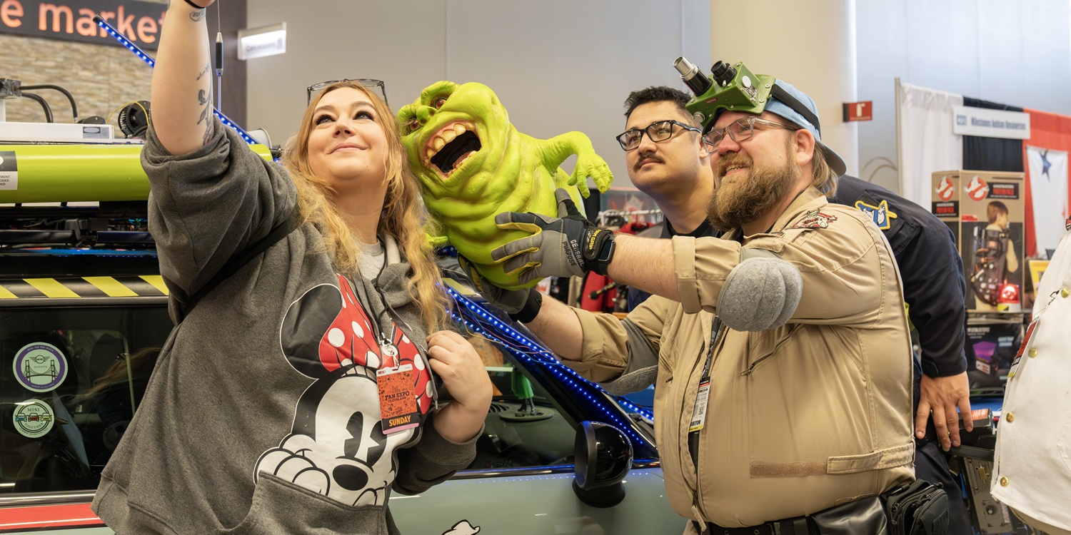 Fans take selfies with a costumed character at FAN EXPO Cleveland. The group smiles and laughs while holding a phone out in front of them, capturing a spontaneous convention moment.