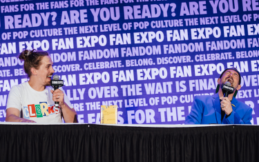 Jason Mewes (left) and Kevin Smith (right) on stage at a table, laughing into their microphones. Jason is wearing a white t-shirt that says "CLERKS" and Kevin is wearing a blue suit jacket with a black t-shirt.