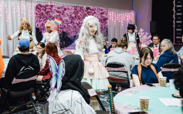 pictured is a programming room where the Maid Café performed. There are multiple round tables with audience members sitting and enjoying their tea. There are white curtains in the background with pink lights and floral backdrops. The Maids are scattered throughout the audience and are entertaining everyone.