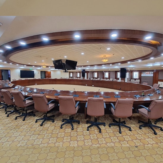 Modern circular boardroom with curved wooden conference table, leather executive chairs, integrated microphones, overhead displays, and elegant coffered ceiling lighting