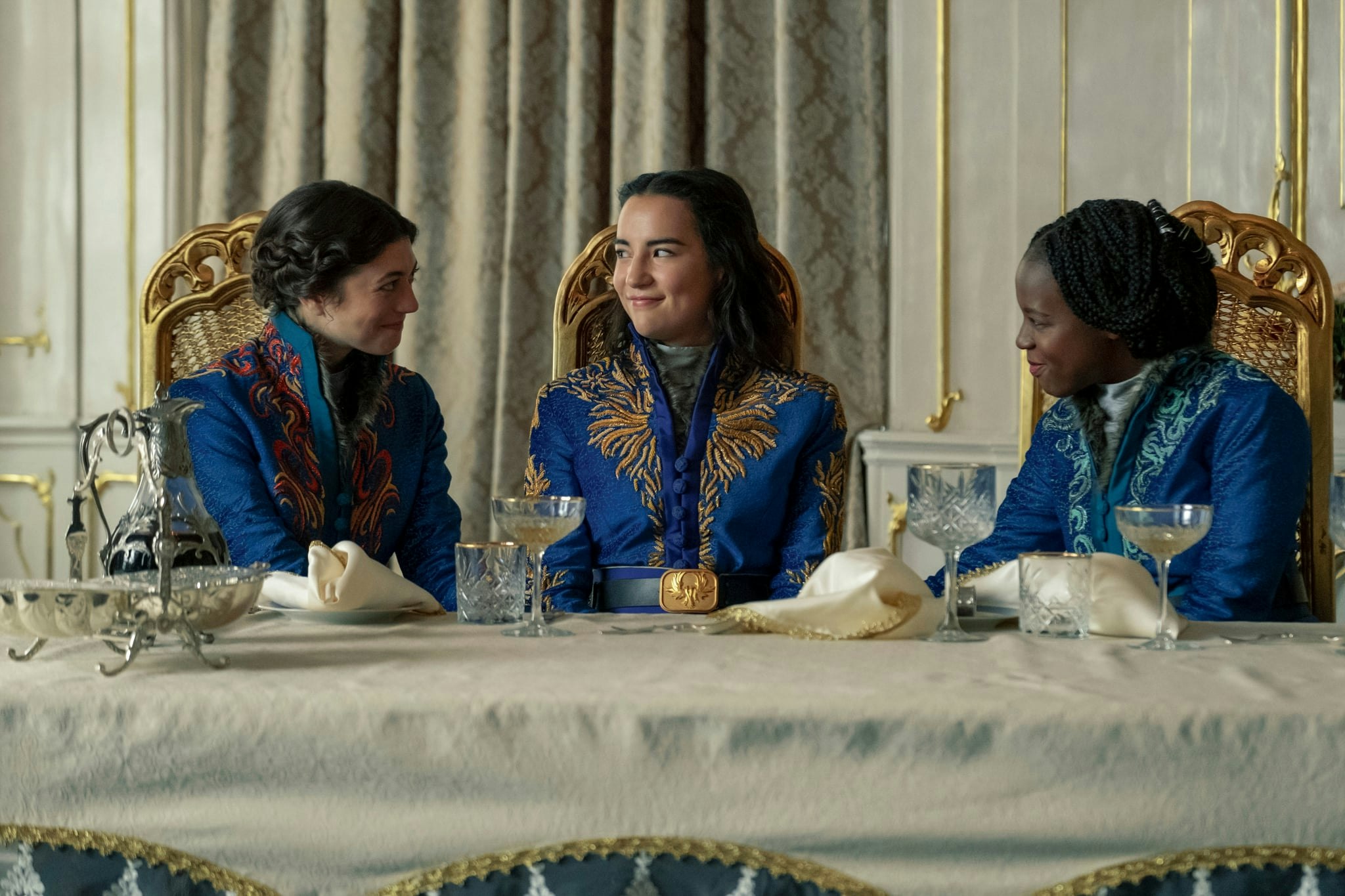 Three characters in very similliar blue and gold jackets sit at a table and smile at each other