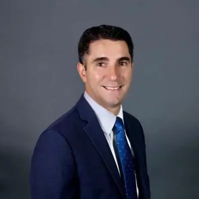Everton Santos, palestrante do setor de saúde, sorrindo e vestindo terno azul com gravata sobre fundo cinza.