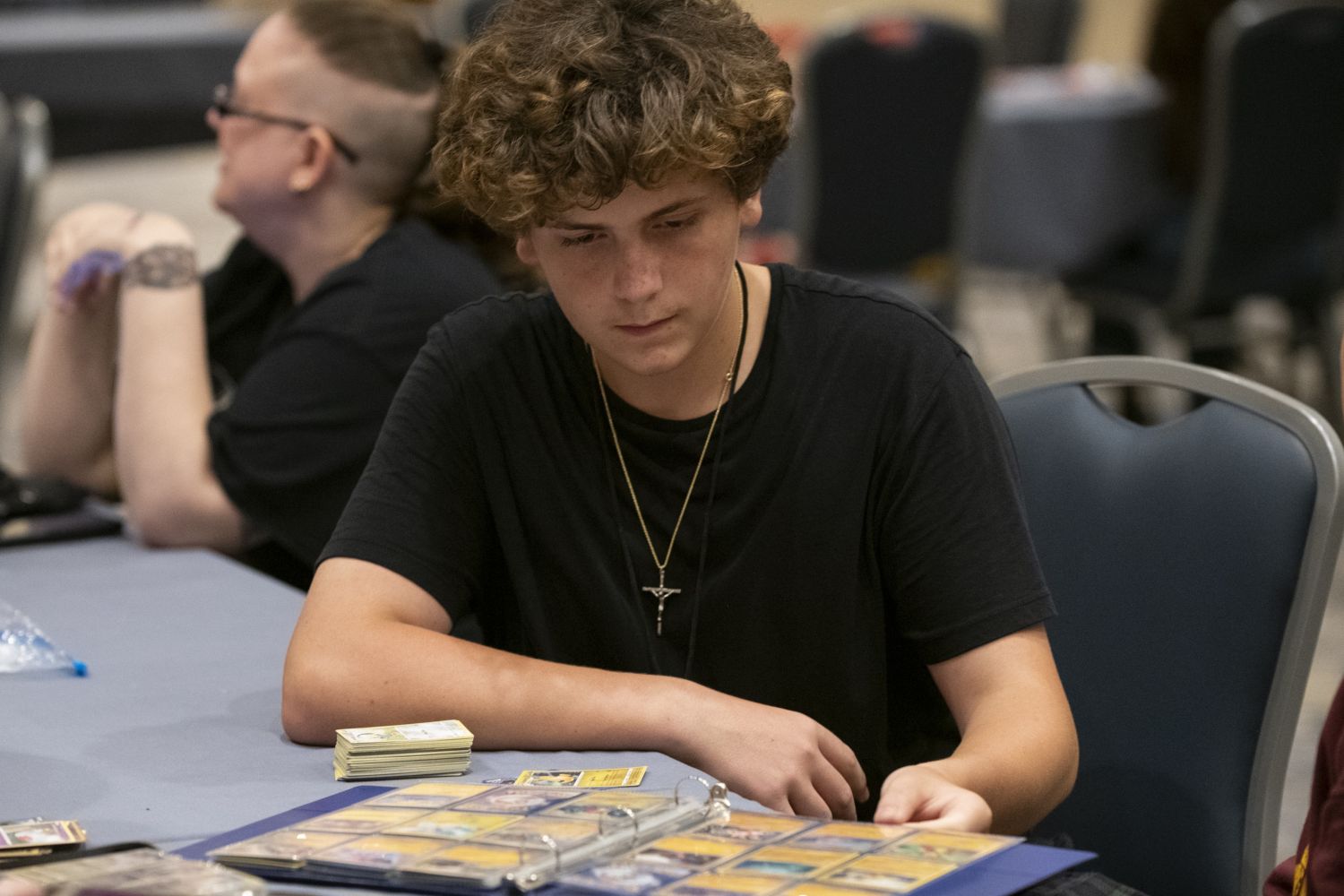 A teenager flips through a binder of Pokémon trading cards while sitting at a table in the gaming area, stacks of cards and binders spread out around him.