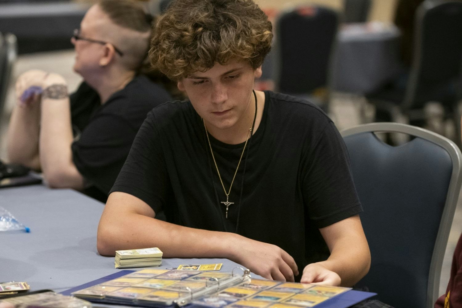 A teenager flips through a binder of Pokémon trading cards while sitting at a table in the gaming area, stacks of cards and binders spread out around him.