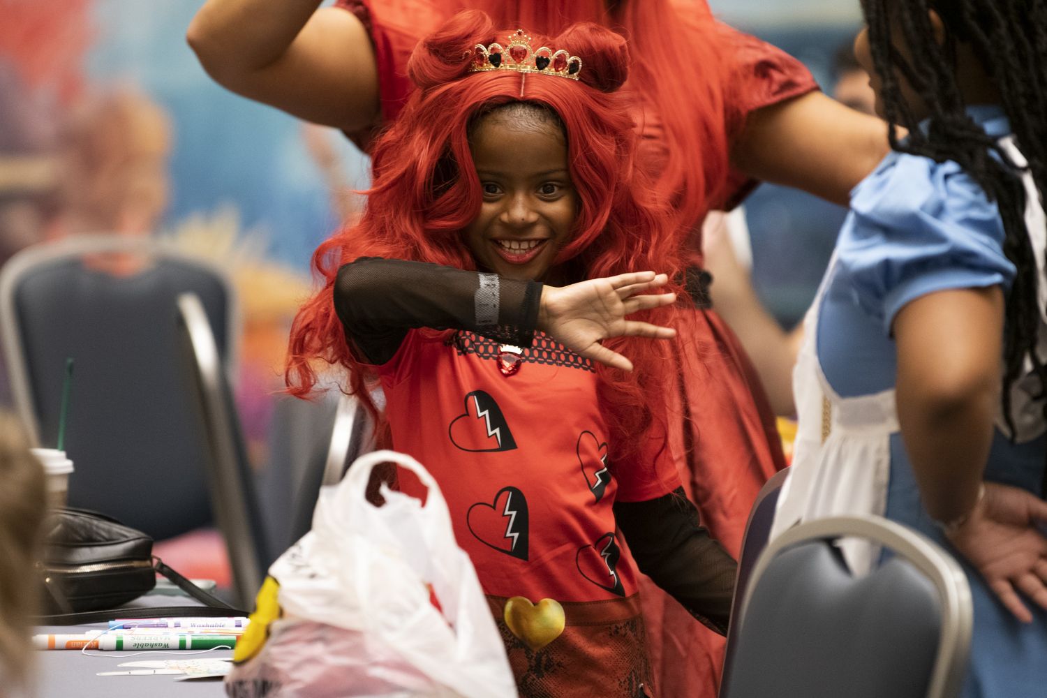 A young girl in a red wig, crown, and costume with broken-heart designs smiles brightly while striking a playful pose in the Kids Zone at FAN EXPO Chicago.