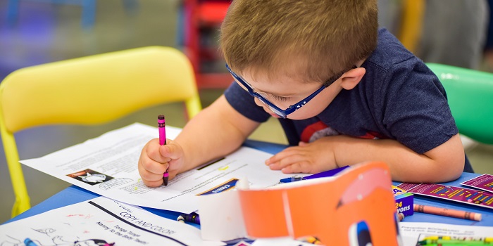 A young child wearing glasses and a navy blue T-shirt is deeply focused on coloring a worksheet with a pink crayon. They are seated at a bright blue table covered with activity sheets, crayons, and a small orange paper craft. Yellow and red chairs surround the table, creating a colorful and inviting space for creative fun.