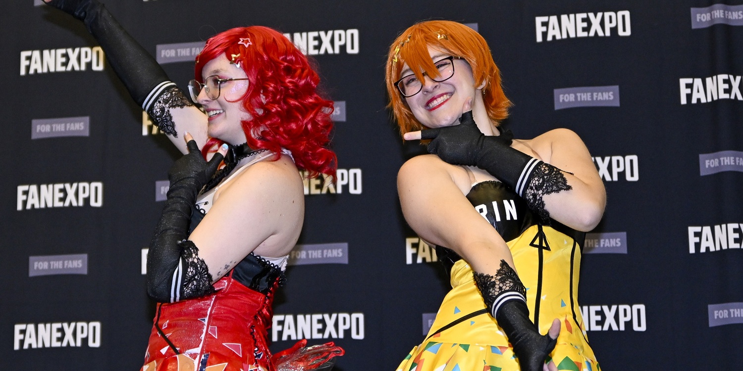 Two cosplayers pose together on a FAN EXPO step-and-repeat backdrop, dressed in colorful anime idol–style outfits. The cosplayer on the left wears a red curly wig, clear glasses, long black lace gloves, and a red and white idol dress with geometric patterns. The cosplayer on the right wears an orange bob wig, glasses, matching black lace gloves, and a yellow idol dress with multicolored accents and “RIN” printed across the top. Both smile brightly and strike energetic idol poses, conveying a fun, performance-ready atmosphere.