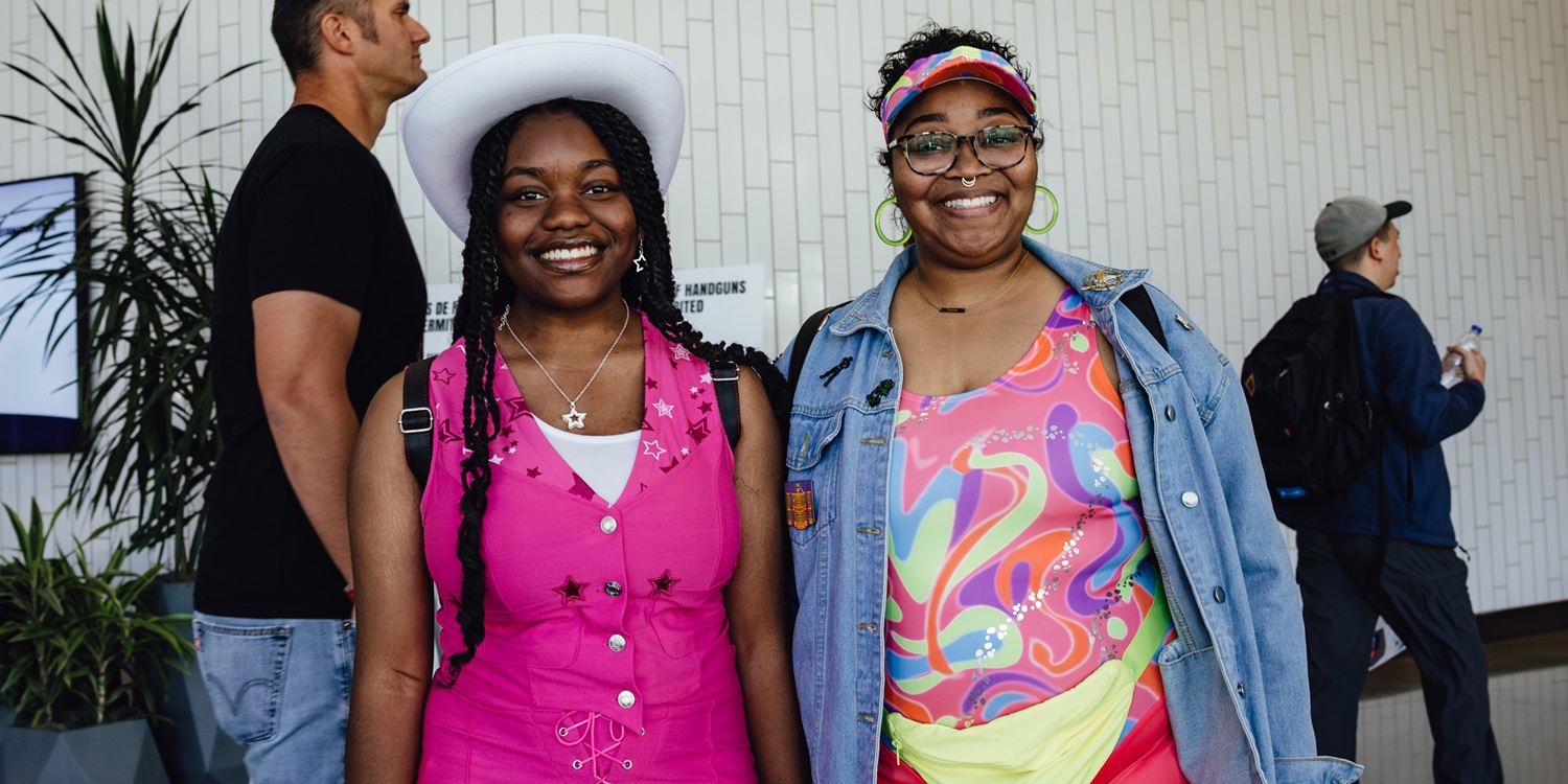 Two friends dressed in colorful Barbie-inspired outfits smile at the camera at Dallas FAN FESTIVAL. One wears a pink vest and white cowboy hat, while the other sports a rainbow-patterned outfit with a denim jacket and visor.