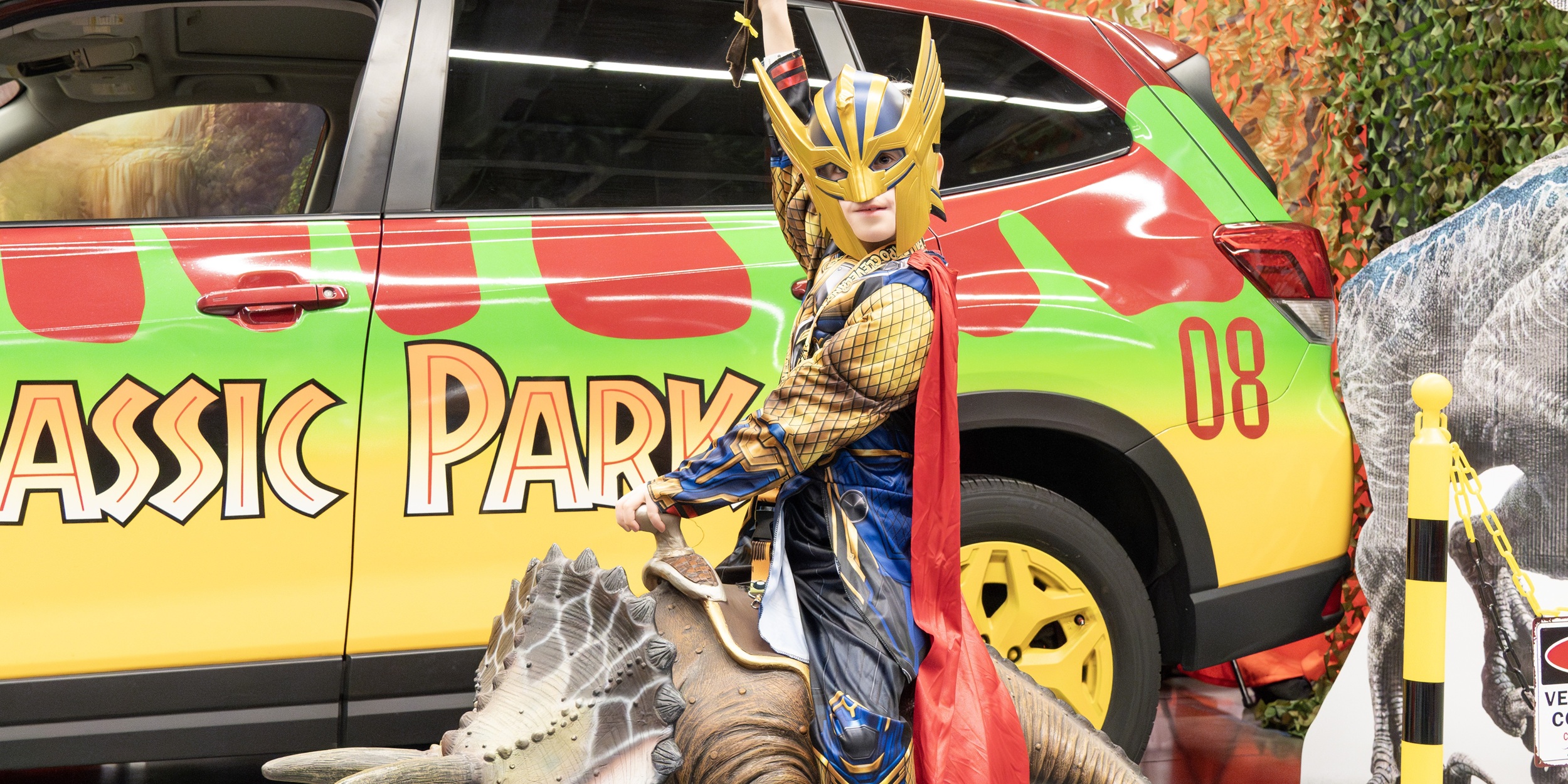A themed vehicle inspired by Jurassic Park is parked indoors with bright green and red graphics. A cosplayer in a horned fantasy costume sits nearby, posing next to the car as part of a fandom vehicle display.