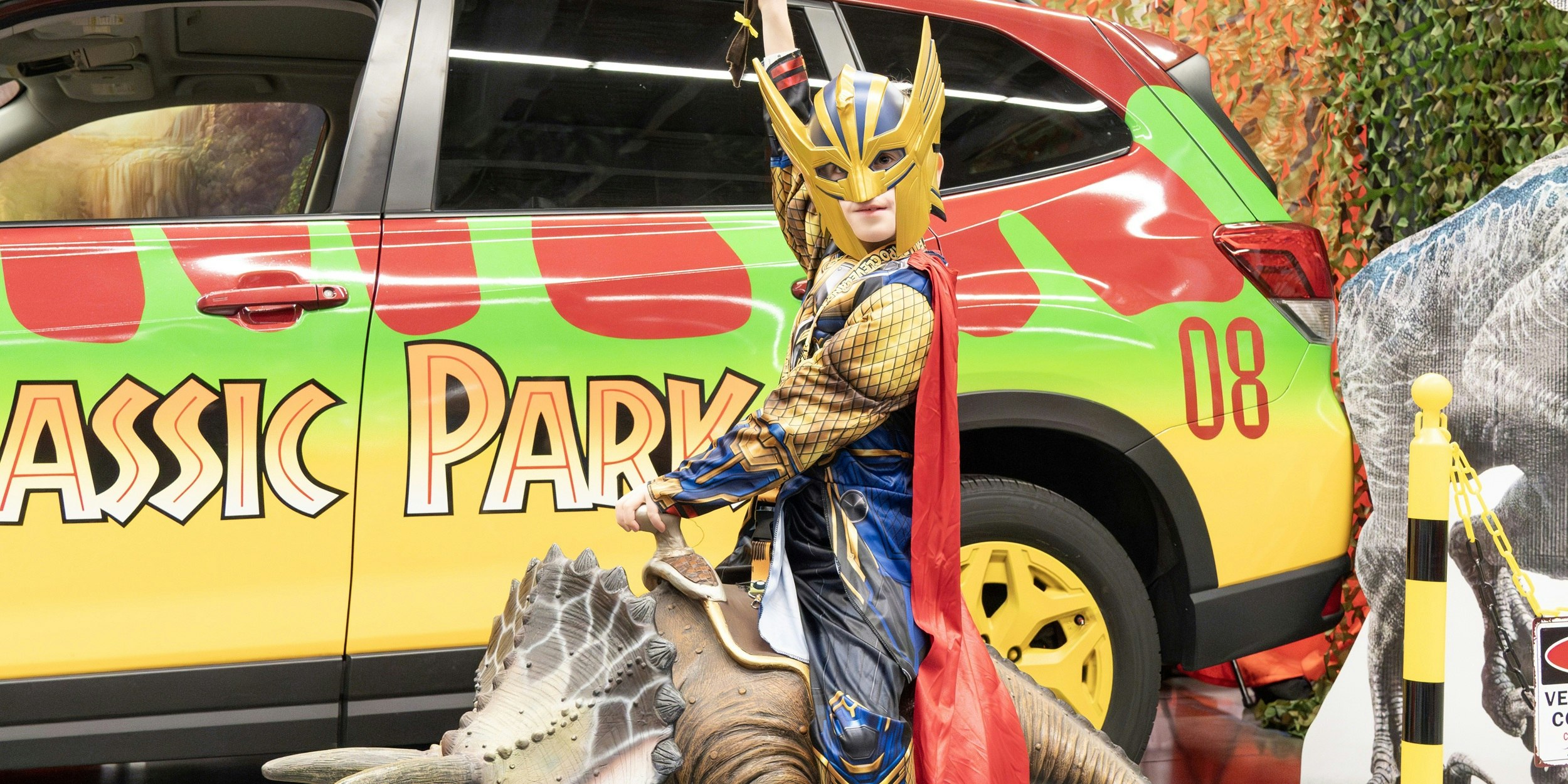 A themed vehicle inspired by Jurassic Park is parked indoors with bright green and red graphics. A cosplayer in a horned fantasy costume sits nearby, posing next to the car as part of a fandom vehicle display.