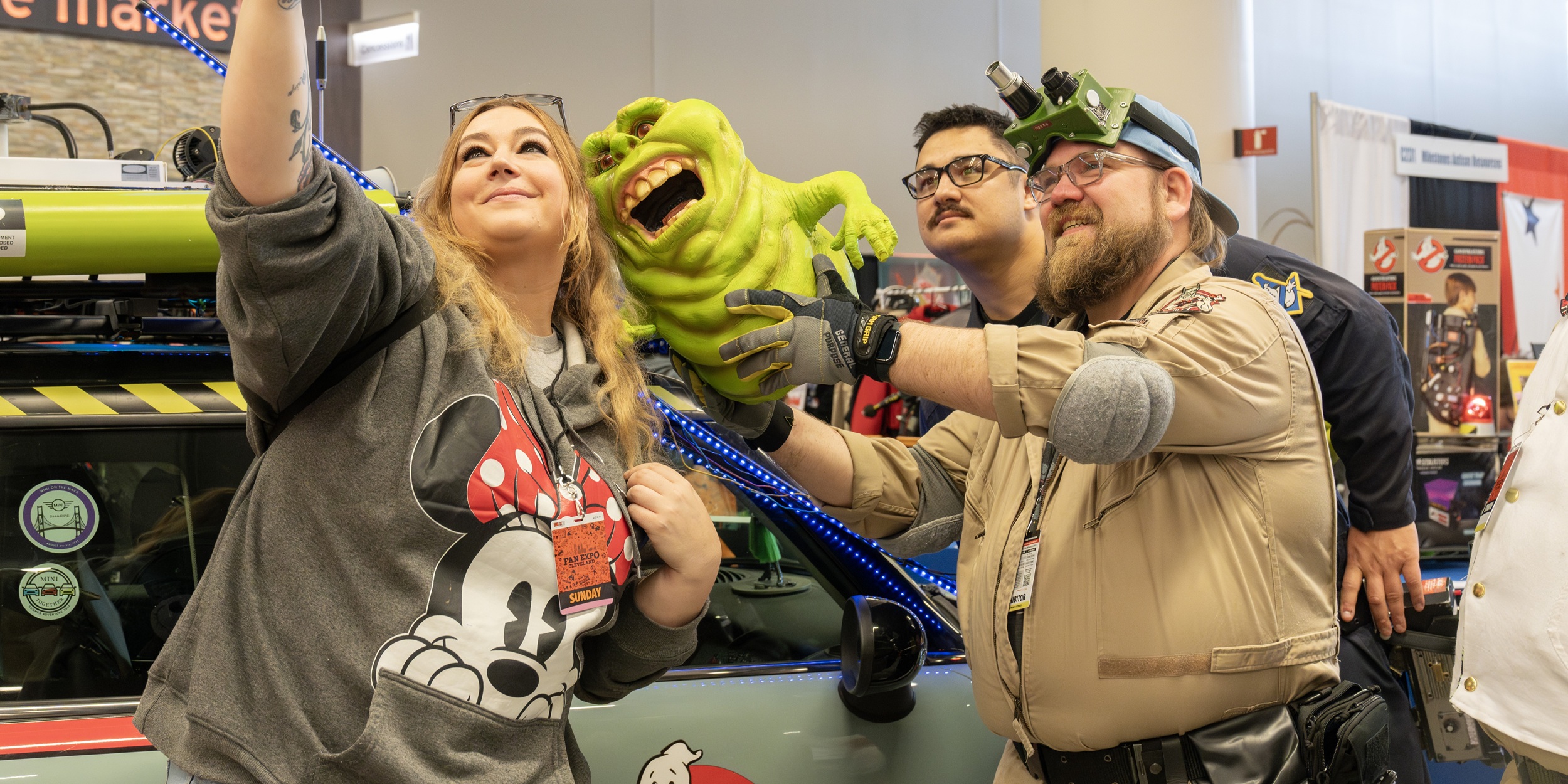 Three fans pose for a selfie with a green ghost puppet at FAN EXPO Cleveland. The group smiles while holding props and wearing themed outfits, capturing a fun moment of fandom connection in the Community Zone.