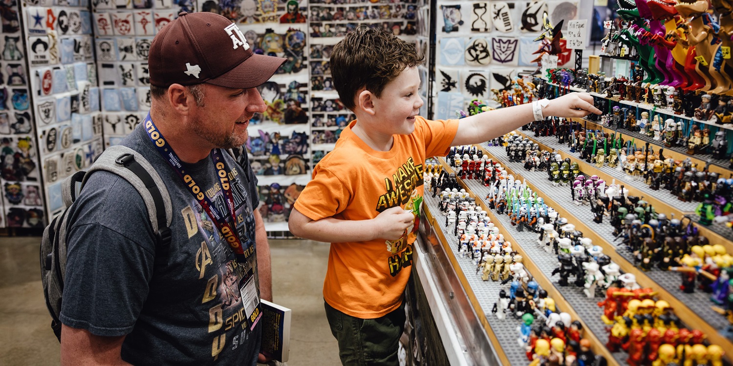 A father and young son browse rows of colorful LEGO minifigures at Dallas FAN FESTIVAL. The boy points excitedly at a figure while his dad smiles beside him, surrounded by walls of pop culture decals.