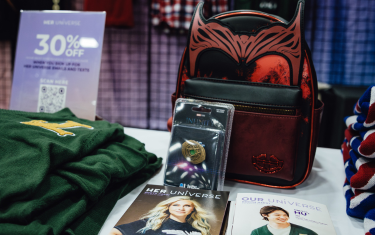 A picture of the Her Universe booth, with a green sweater on the left, a poster advertising 30% off the merchandise, a red WandaVision backpack, a pin, and some brochures on the table.