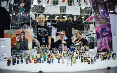 Three women sitting at their booth, selling animated print art, as well as little figurines.