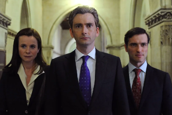 David Tennant in a suit with a blue tie standing in between a woman (in black blazer and white blouse) and a man (black suit and red tie) as they walk down a marble hallway