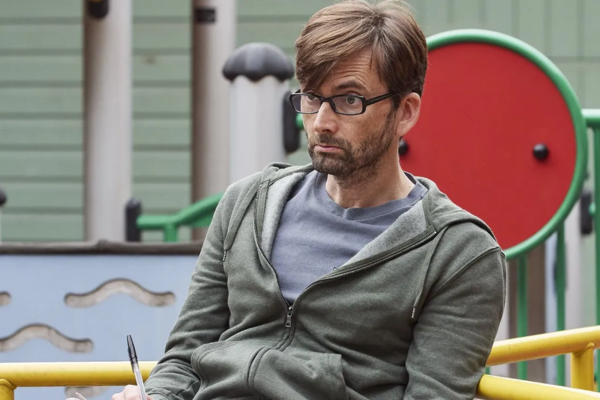 Looking a tad disheveled and in a green gray hoodie and gray tshirt, David Tennant is sitting at a playground