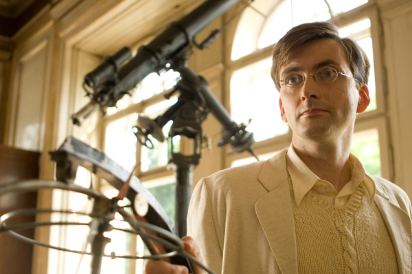Standing next to a telescope, David Tennant is wearing an all white ensemble (white jacket, white sweater and a white button up.