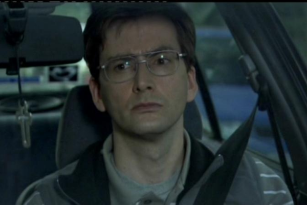 In old school 70's looking wired glasses, he is sitting in a car and looking off to the side