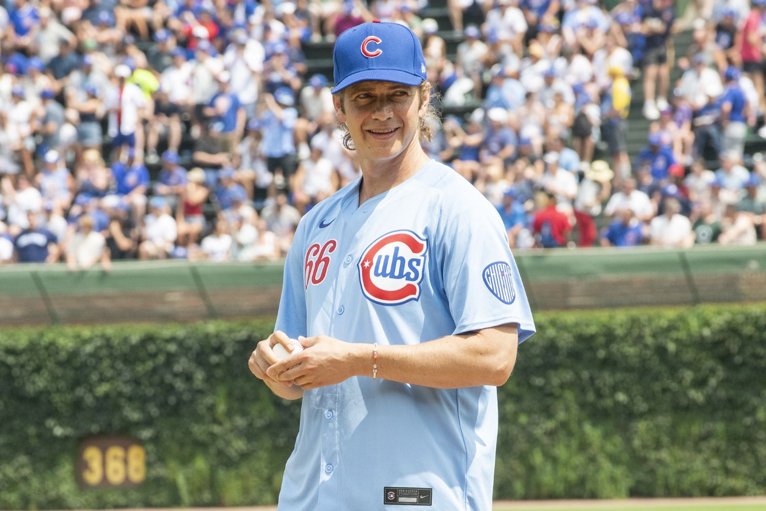 Hayden Christensen stands on the pitcher’s mound at Wrigley Field, smiling in a light blue Cubs jersey and cap while holding a baseball. The crowd fills the stands behind him.