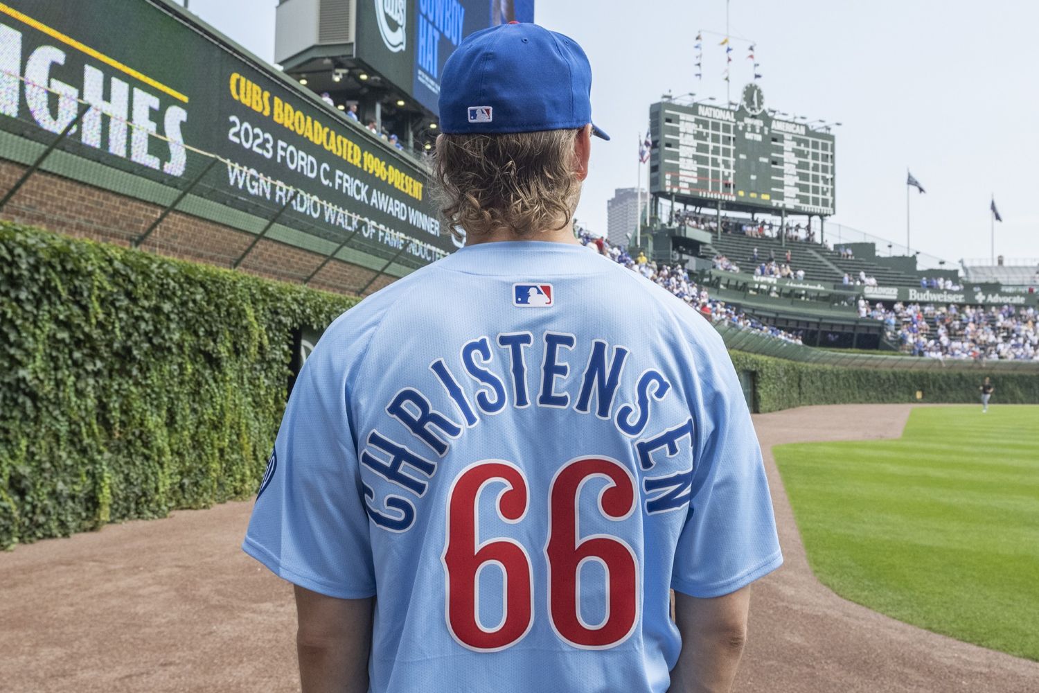 From behind, Hayden Christensen looks out at the field and scoreboard while wearing a Cubs jersey with his name and the number 66 across the back.