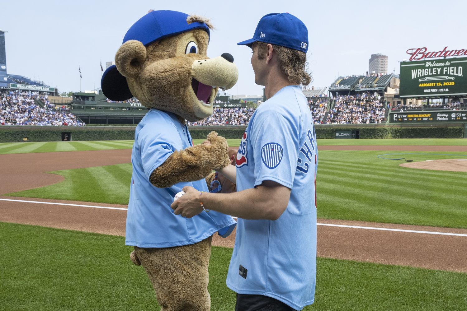 Hayden Christensen shakes hands with Clark the Cub mascot near the field, both wearing Cubs jerseys as the stadium crowd looks on.