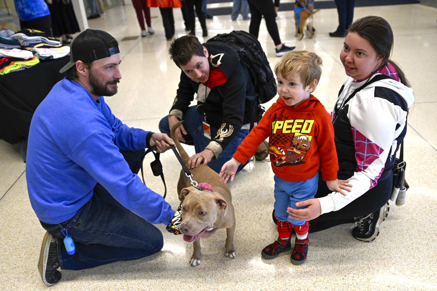 A young boy reaches out in delight to pet a calm, friendly dog while being supported by a woman in a white Spider-Gwen hoodie. A man in a blue sweater kneels to hold the dog steady, while another attendee crouches nearby, watching the heartwarming moment unfold.
