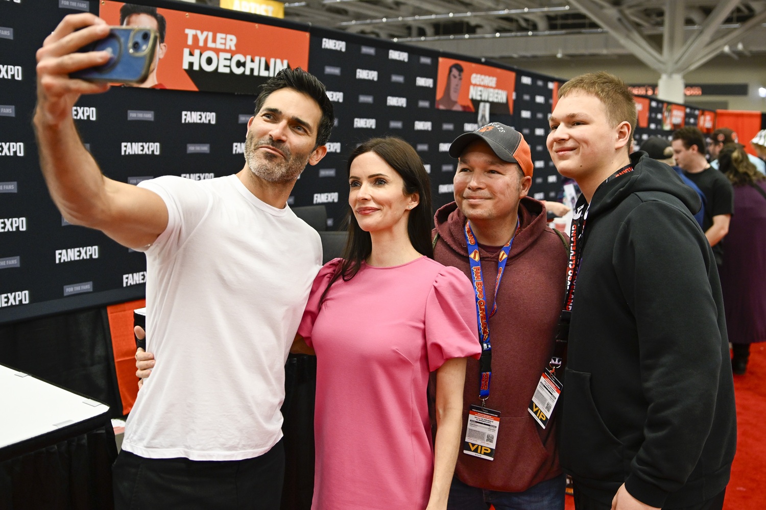Tyler Hoechlin, dressed casually in a white T-shirt, holds out a phone for a selfie with a smiling father and son at his booth. His co-star, Bitsie Tulloch, stands beside him in a bright pink dress, while the fans grin widely with VIP badges around their necks.