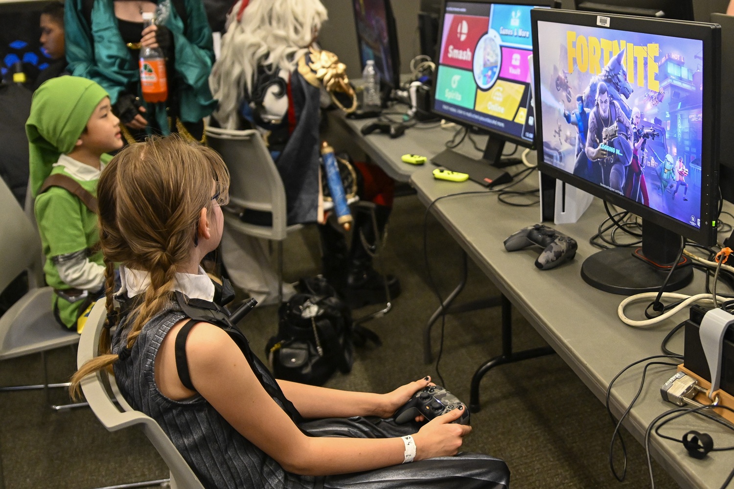 A girl in costume, possibly cosplaying a stealthy or tactical character, sits in a gaming lounge at FAN EXPO. She’s focused intently on a monitor displaying Fortnite as she grips a controller. Other costumed gamers, including one dressed as Link, play alongside her in the background.