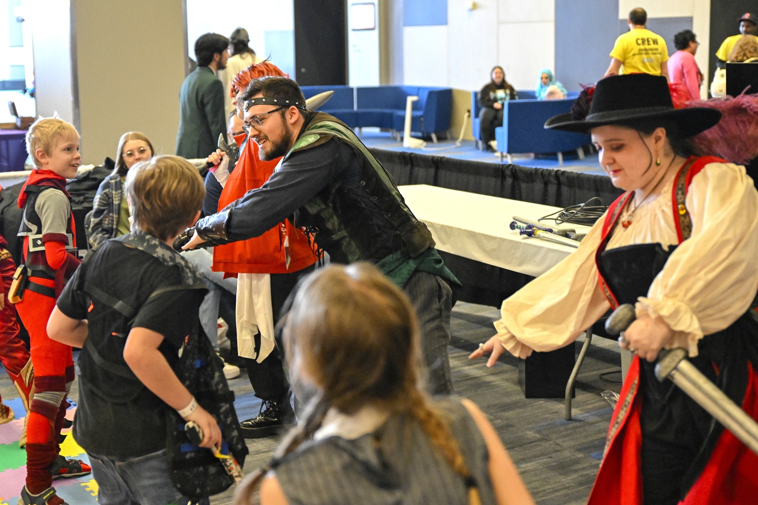 A group of children gathers around two costumed performers dressed as fantasy adventurers—one in green leather armor wielding foam weapons, and the other in a red and black swashbuckler outfit with a wide-brimmed hat. The children eagerly engage in the playful duel, wide-eyed and smiling in excitement.
