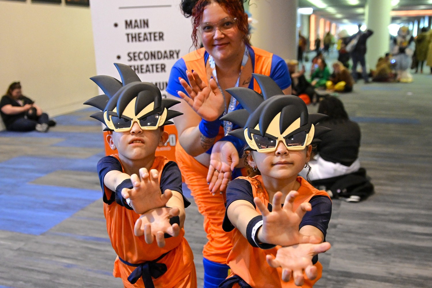 Two young kids dressed as Goku from Dragon Ball Z, complete with orange training outfits and oversized Goku hair sunglasses, strike a dramatic Kamehameha pose. Behind them, an adult in matching cosplay playfully mimics the stance, grinning proudly as the trio poses in perfect anime sync.
