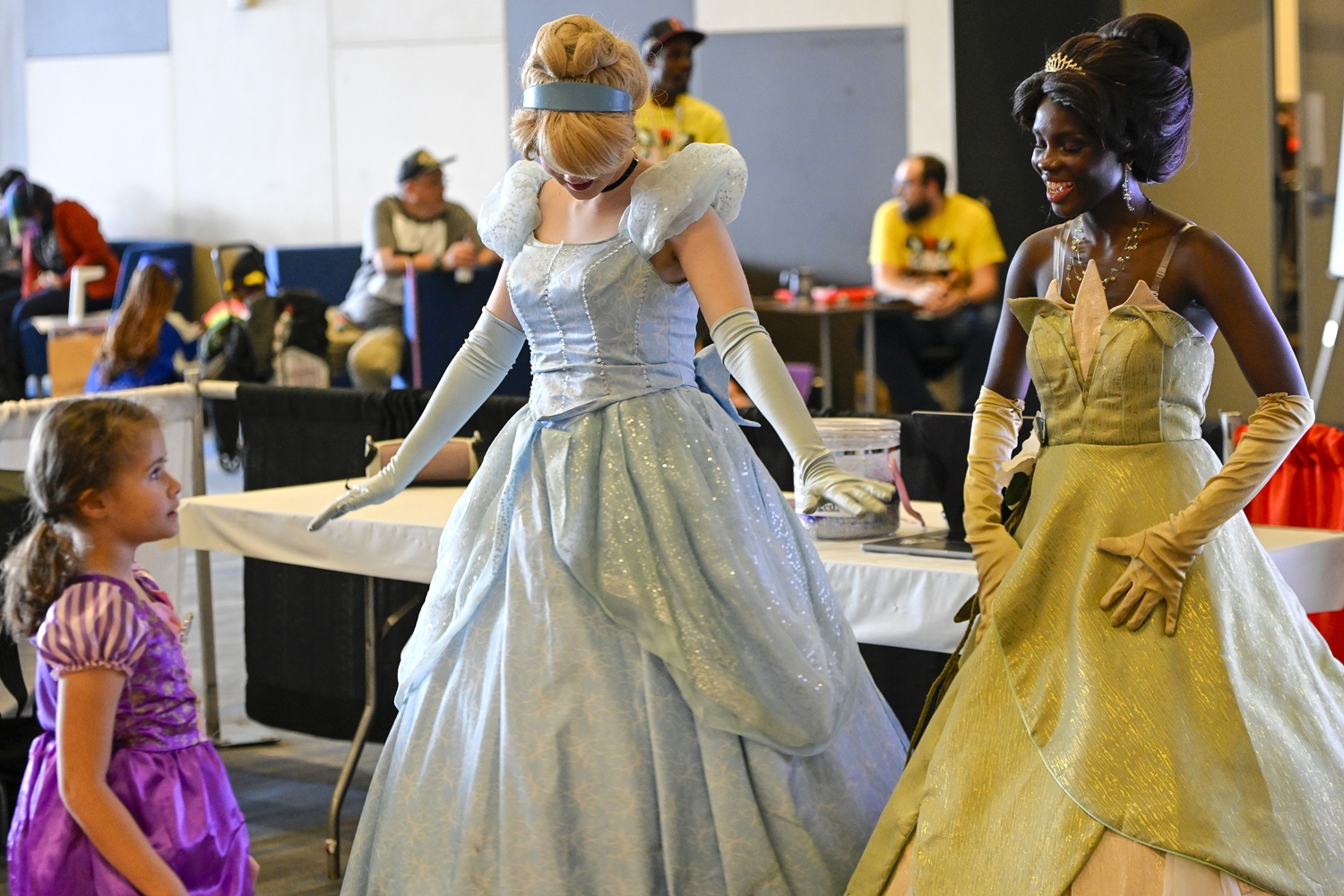 A little girl in a Rapunzel dress beams up at two elegant cosplayers portraying Cinderella and Princess Tiana. Cinderella curtsies gracefully, while Tiana offers a warm smile, making the magical princess moment feel straight out of a fairy tale.