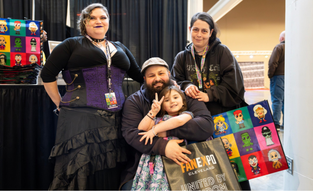 The image depicts a heartwarming family moment at FAN EXPO Cleveland. Two adults and one child are posing together with genuine smiles. The man, seated, embraces a young girl who is playfully making a peace sign. The woman stands behind, all dressed in casual, fan-inspired attire. The child holds a bag featuring colorful character artwork, suggesting their shared interest in pop culture. Behind them, a display booth with vibrant and stylized character portraits adds to the festive atmosphere of the event. This scene captures the family-friendly nature of the expo, emphasizing that such events are enjoyed by fans of all ages.