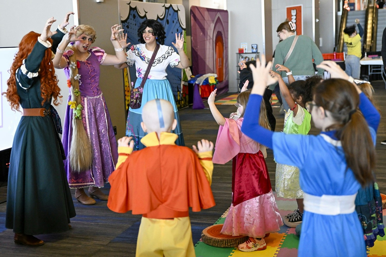 Cosplayers dressed as Merida, Rapunzel, and Mirabel from Disney films lead a group of children through an interactive activity. The kids, also dressed as various animated characters, follow their lead with raised arms, fully immersed in the moment of pretend play and movement.
