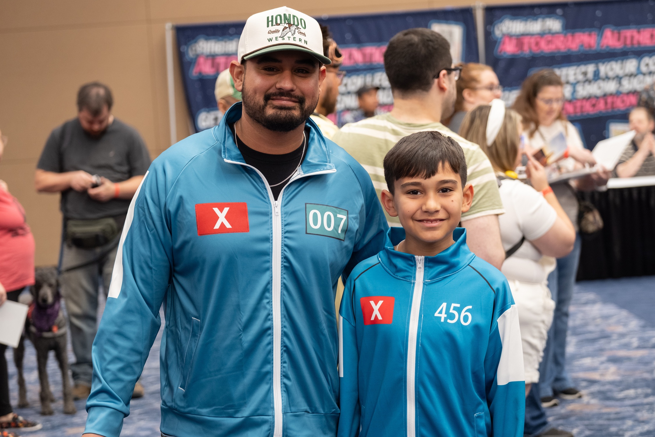 A father and son dressed in matching teal tracksuits inspired by Squid Game smile while standing in line inside the show floor.