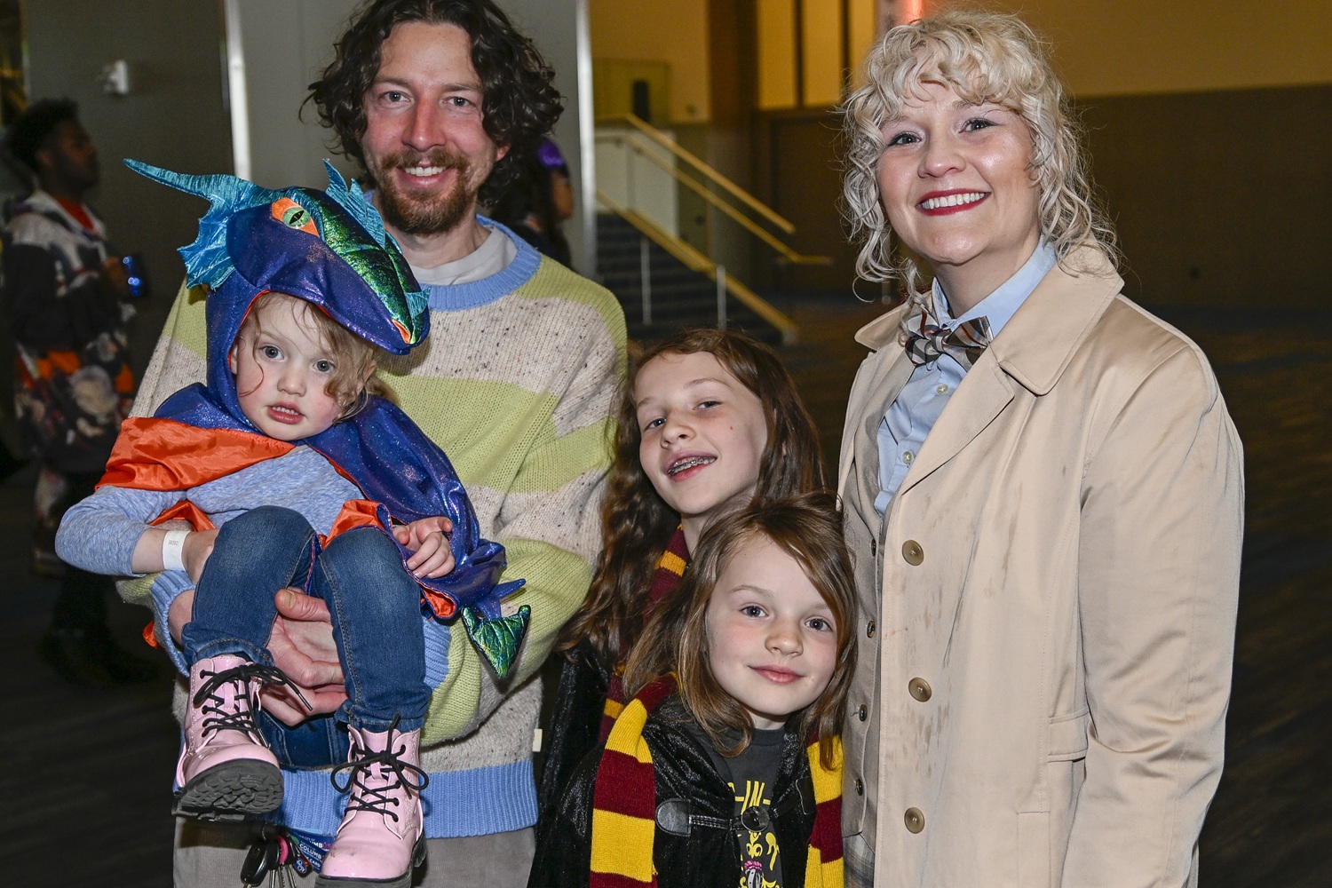 A smiling family of five poses together. A young child dressed as a shiny blue-and-orange dragon is perched in their father’s arms. The rest of the family beams at the camera, with one child in a Gryffindor scarf and another in a Doctor Who-inspired trench coat and bowtie.