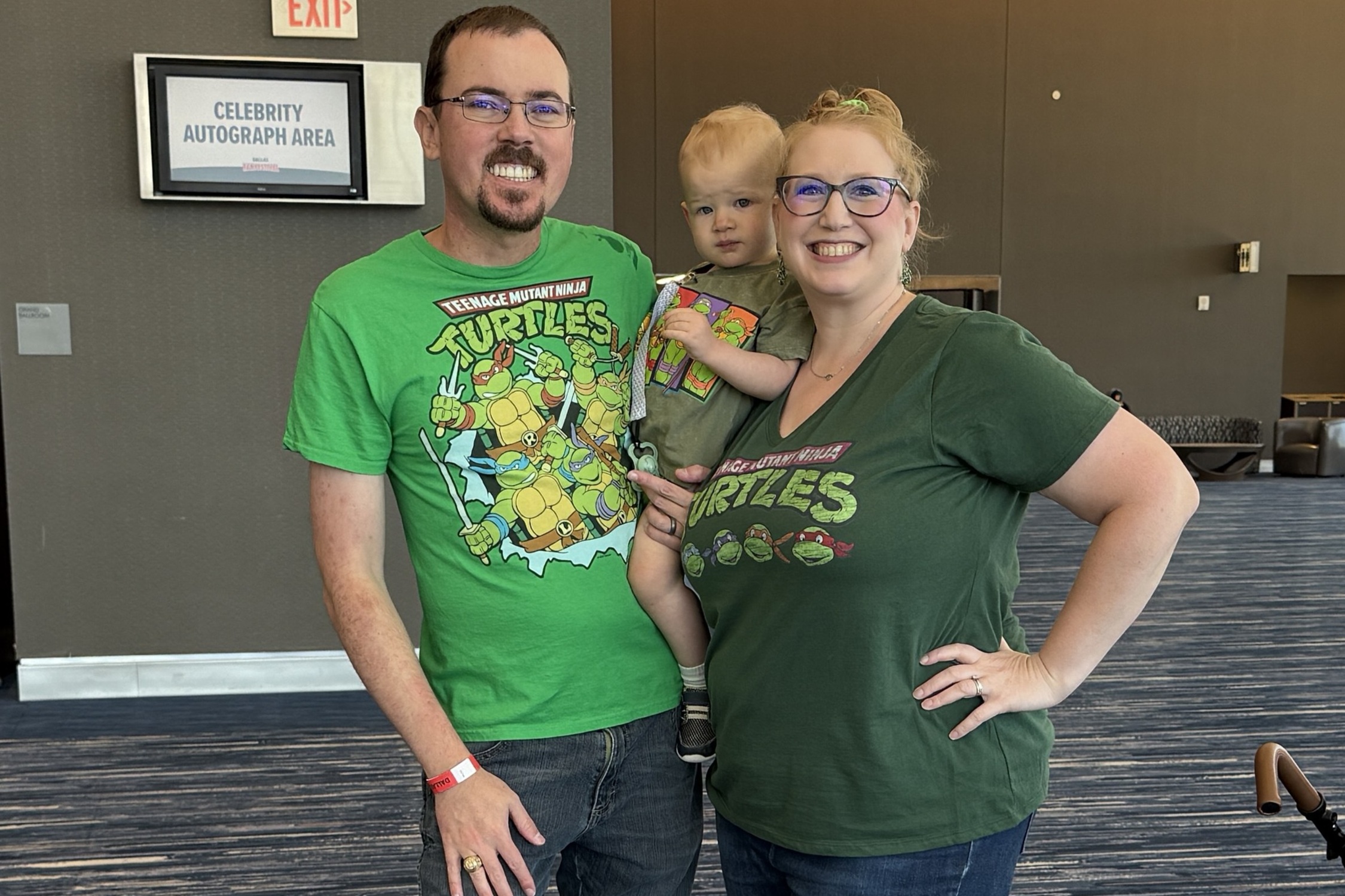 A smiling family of three dressed in matching Teenage Mutant Ninja Turtles shirts pose together near the Celebrity Autograph Area, holding their young child.