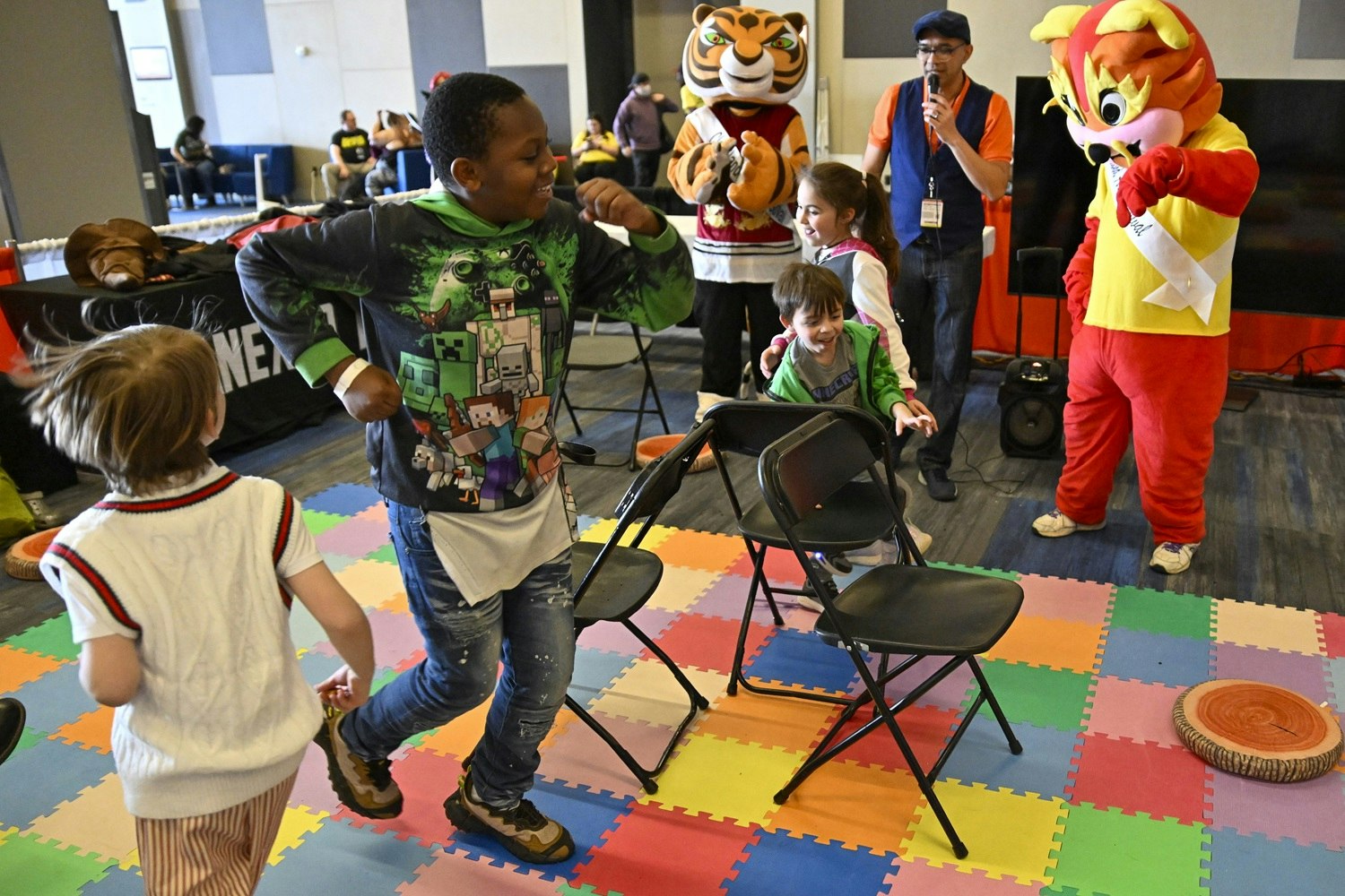 Children dash around a ring of chairs during a fast-paced game of musical chairs. In the background, two cheerful tiger mascots clap and dance along. A staff member with a mic energizes the game, while kids laugh and scramble to claim a seat before the music stops.