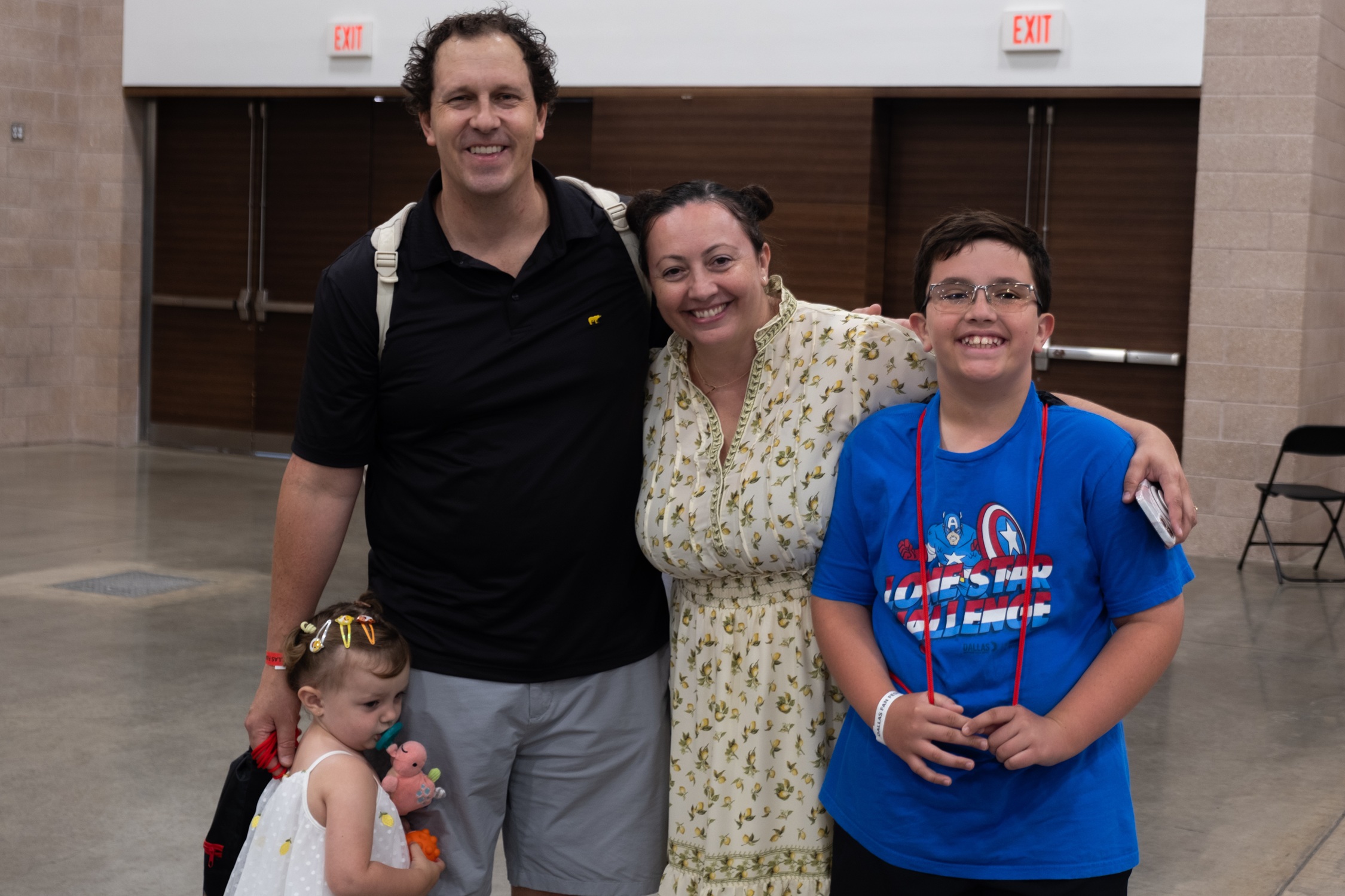 A smiling family of four poses inside the convention center — a man, woman, young boy in a Captain America shirt, and a toddler holding a plush toy.