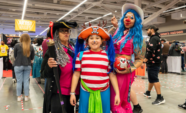 The image features a vibrant and colorful family dressed in playful costumes at FAN EXPO. On the left, an adult in a pirate costume holds a faux sword, wearing a black hat with a skull design. The child in the center is dressed in a colorful, striped outfit reminiscent of a whimsical character, complete with a rainbow tutu and a playful hat. To the right, another adult wears a bright pink and blue outfit, adorned with a large pirate hat and holding a soccer ball. Their cheerful expressions and the imaginative costumes create a festive and fun atmosphere, illustrating the family-oriented and creative spirit of the event. The background shows the bustling environment of the expo, with other attendees and booths, enhancing the lively setting.
