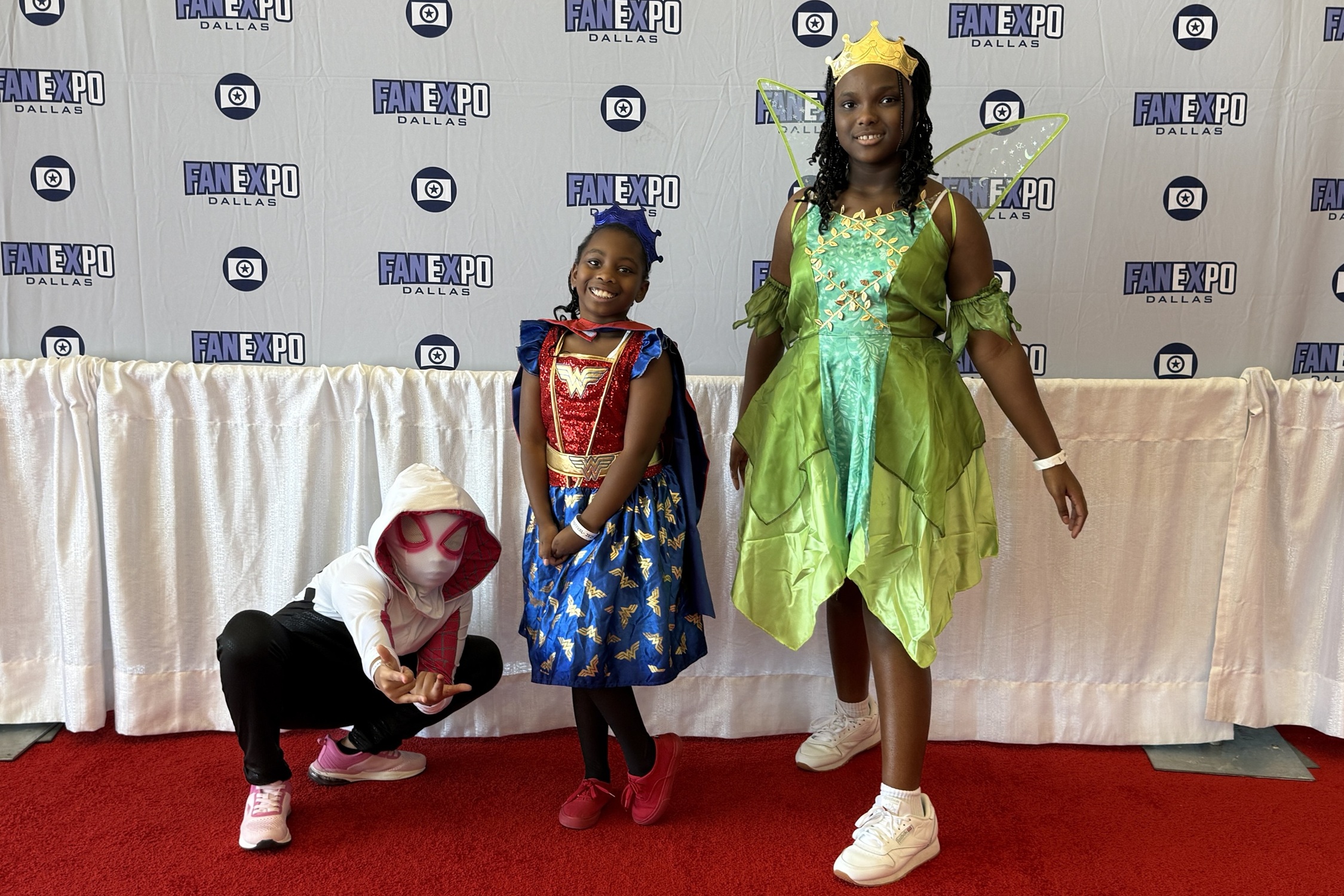 Three children pose on the red carpet in front of a FAN EXPO Dallas backdrop, dressed as Spider-Gwen, Wonder Woman, and Princess Tiana.