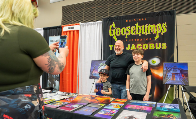 The image captures a cheerful moment at a convention booth for "Goosebumps," featuring the original illustrator, Tim Jacobus. He stands between two young fans, smiling broadly as they pose for a photo taken by another attendee visible in the foreground. The booth is adorned with vibrant and eerie artwork from the "Goosebumps" series, displayed on the table and the banner behind them. This engaging scene showcases the interaction between fans and creators, highlighting the personal connections and memorable experiences that fan conventions like FAN EXPO provide.