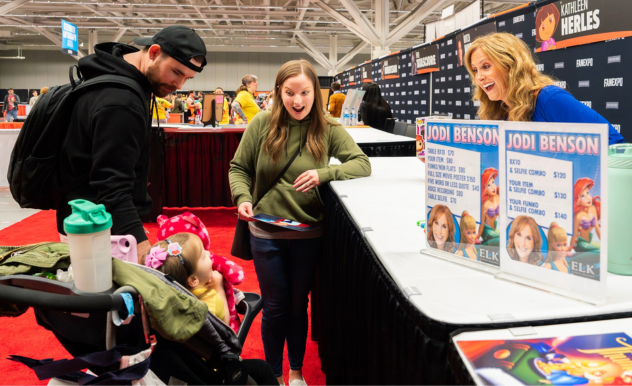 The image captures a family interacting with voice actress Jodi Benson at a FAN EXPO event. The scene shows a young couple and their small child, who is sitting in a stroller, engaging with Jodi, who is animatedly responding with a joyful expression. The father, wearing a black beanie and backpack, looks on while the mother, visibly excited, talks to Jodi, holding an item for autographing. Jodi Benson’s booth is decorated with signs featuring her name and photos, creating a personal and engaging atmosphere for the fans. This image beautifully illustrates the memorable interactions that can occur at fan conventions, bridging the gap between celebrities and attendees.
